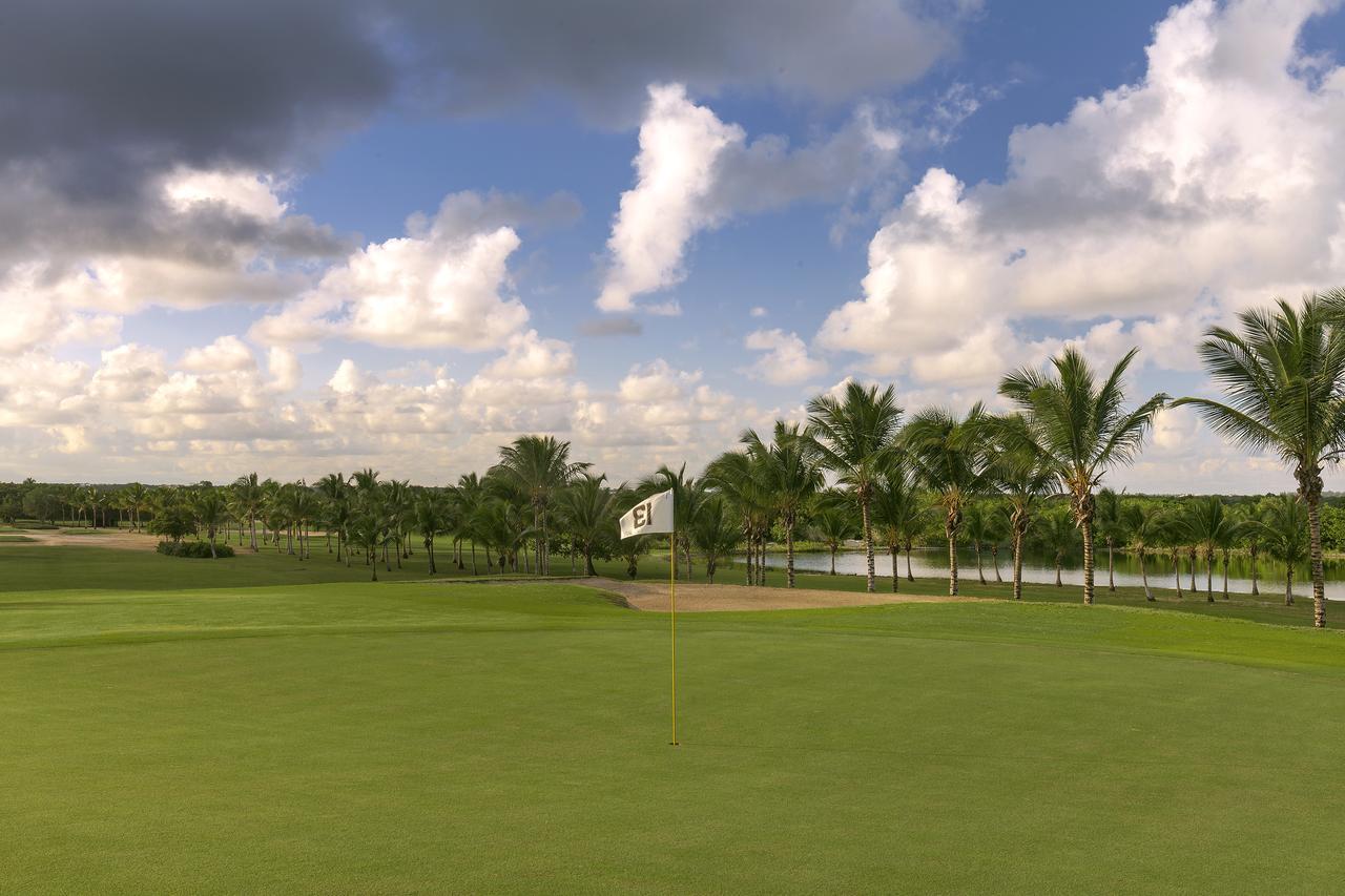 Catalonia Punta Cana Exterior foto The photo shows a scenic view of a golf course. In the foreground, there is a well-maintained green area with a flag indicating the location of the hole. Surrounding the green are clusters of palm trees, suggesting a tropical or warm climate. In the 