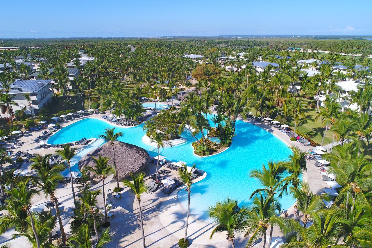 Catalonia Punta Cana Exterior foto The photo shows a beautiful aerial view of a tropical resort featuring a large, winding swimming pool surrounded by palm trees. The pool has several islands and small areas with loungers for relaxation. The surrounding landscape is lush and green, wi