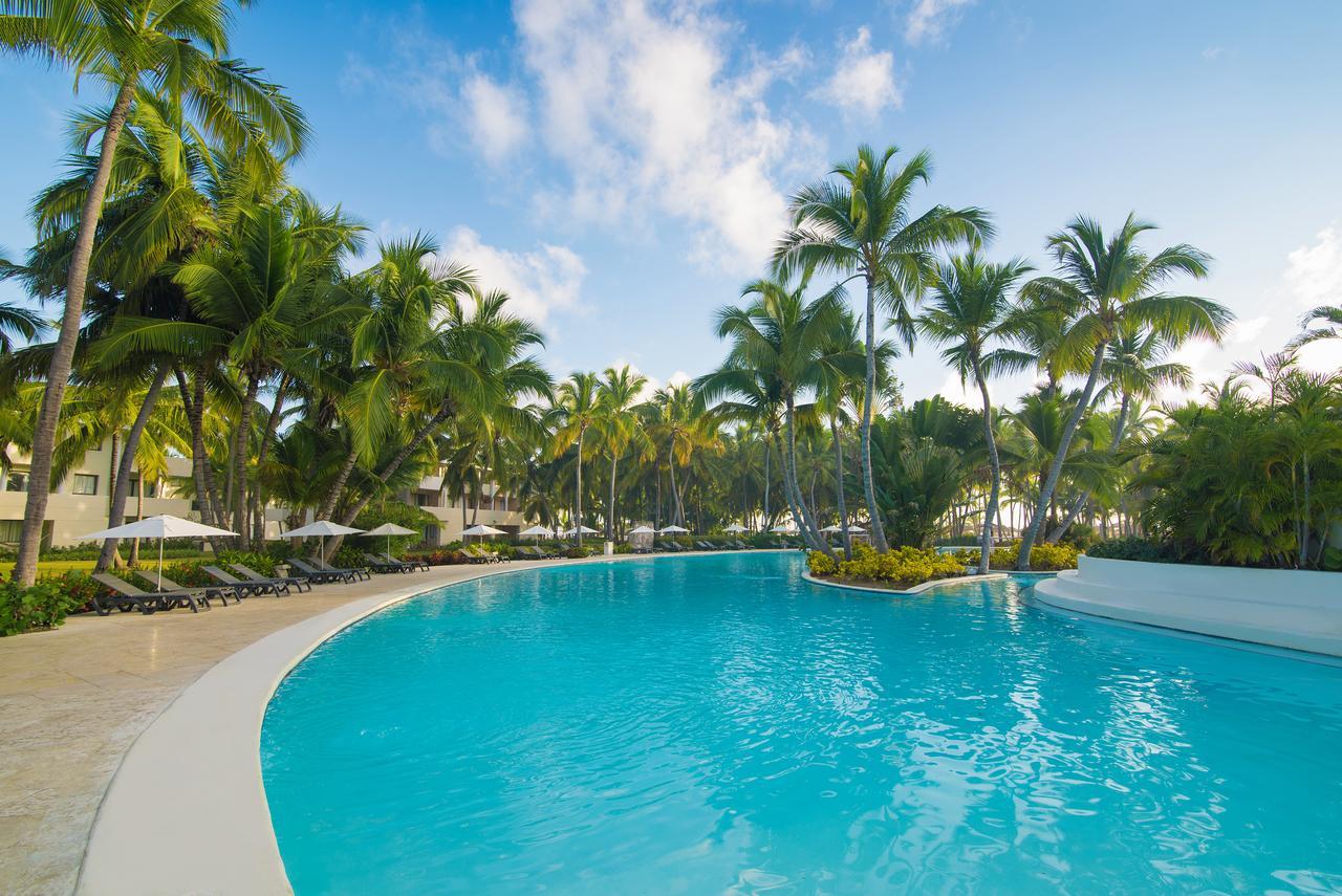 Catalonia Punta Cana Exterior foto The photo shows a beautiful resort scene featuring a large, inviting swimming pool with a curved shape. Surrounding the pool are lush palm trees, creating a tropical atmosphere. The sky is bright and clear, indicating a sunny day. Comfortable lounge 